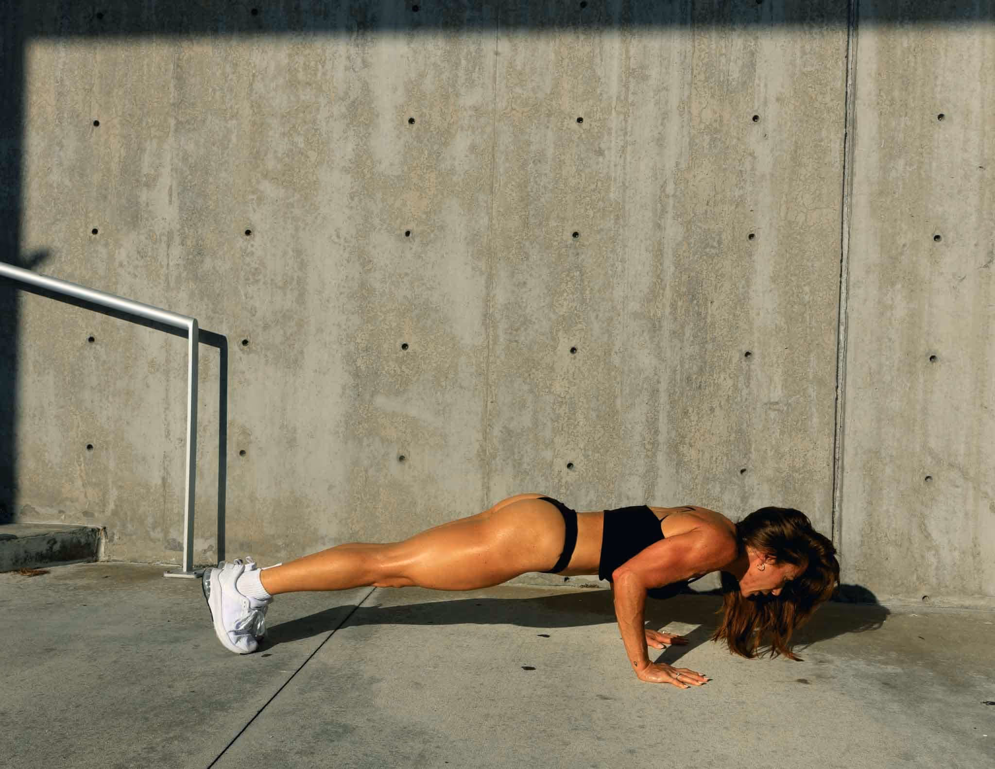 Selena in a low push-up position on sunlit concrete beside a pale wall