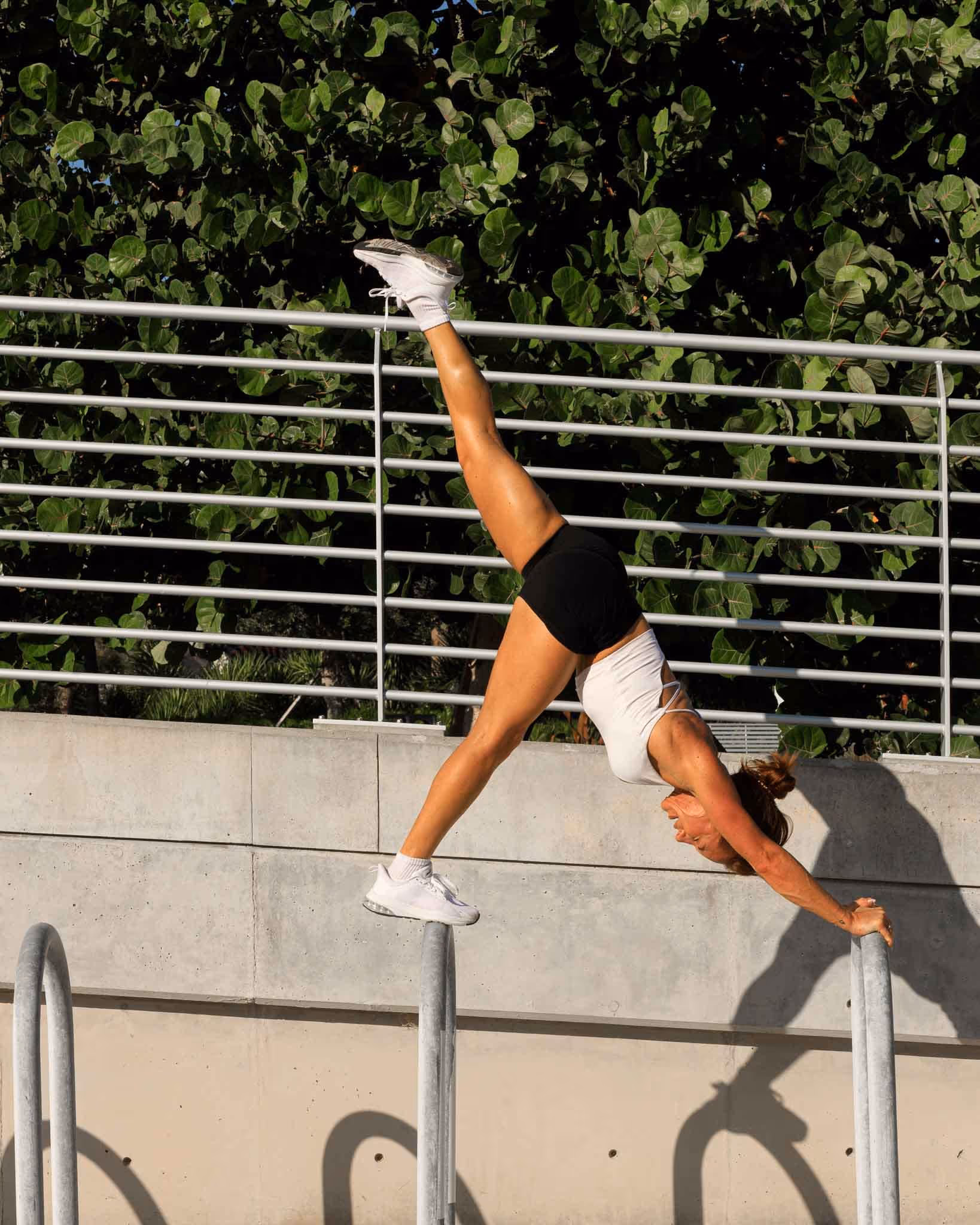 Selena holding a handstand on a metal rail in bright sunlight, legs split in a wide straddle